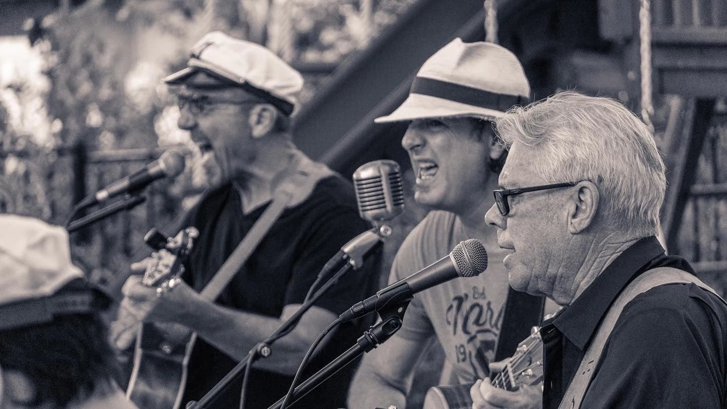 Three musicians performing into microphones, with two playing guitars.